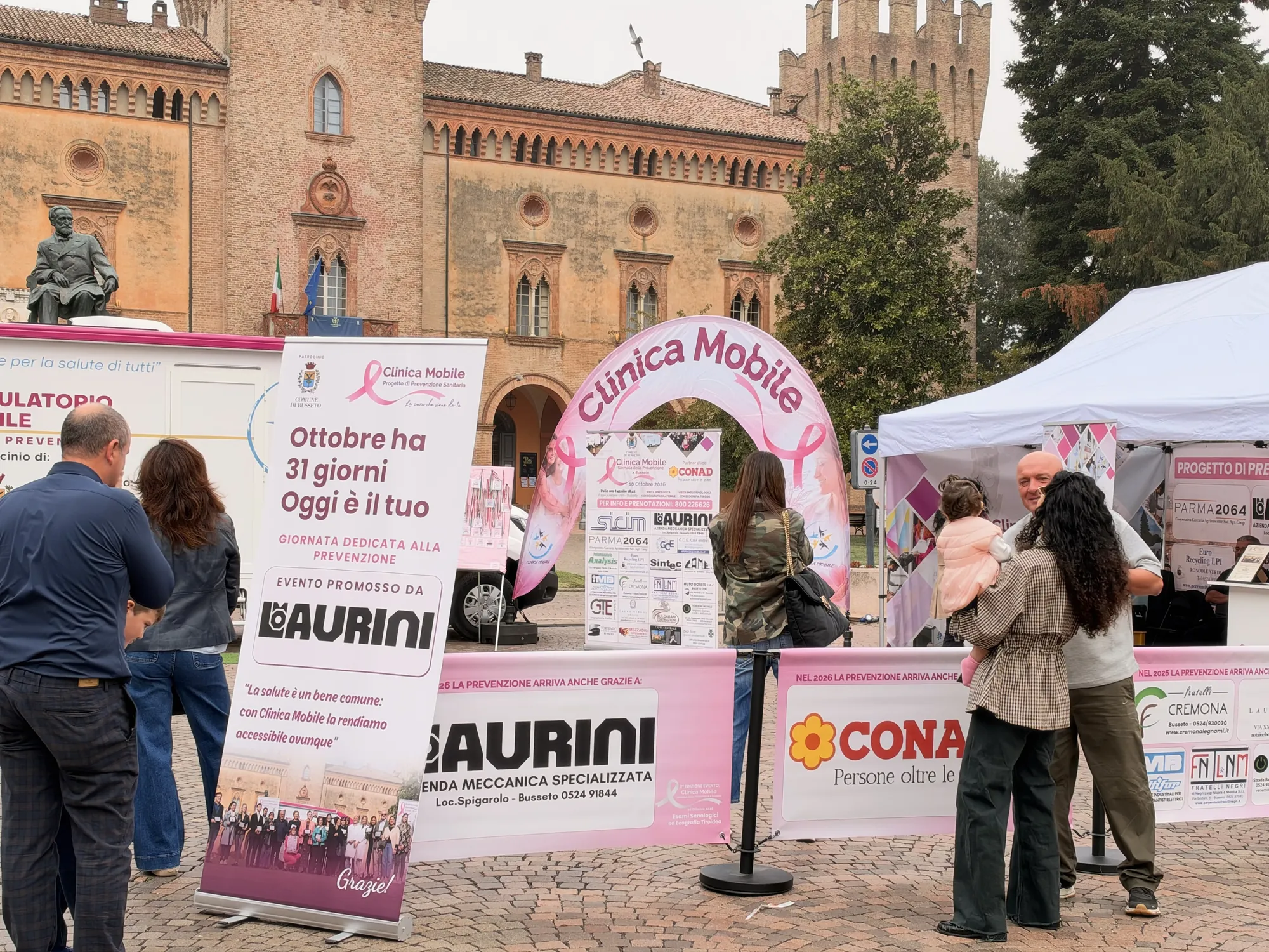 Piazza Verdi a Busseto piena di persone davanti all'ambulatorio mobile e all'arco rosa di Clinica Mobile